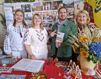 Trois bénévoles en chemise brodée et un visiteur tenant un dépliant, derrière la table du stand